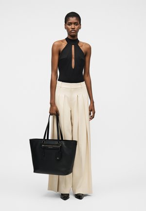 Vrouw met een zwarte haltertop en wijde beige broek die een grote zwarte draagtas vasthoudt, staand tegen een effen achtergrond.