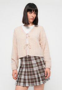 Cardigan court rose clair avec un design en tricot texturé et une fermeture à nouer, assorti d'une jupe plissée à carreaux marron et blanc.