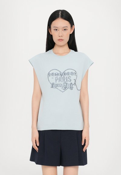 T-shirt bleu clair à manches courtes avec un graphique de cœur et le texte "QUELQU'UN À PARIS M'AIME" dans une couleur contrastante. Tissu doux, coupe décontractée.