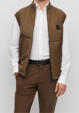 Homme portant une veste matelassée sans manches marron sur une chemise blanche habillée, associée à un pantalon marron et une ceinture noire, les mains dans les poches.