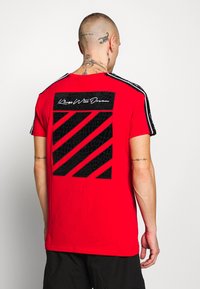 Camiseta roja de manga corta con un diseño gráfico texturizado en negro en la parte trasera que presenta rayas diagonales y el texto "Kings Will Dream".