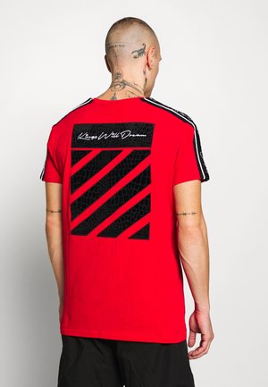 Červené tričko s krátkým rukávem, na zadní straně s texturovaným černým grafickým designem, který obsahuje diagonální pruhy a text "Kings Will Dream".