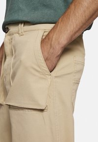 Beige cargoshorts med sidelommer, bæltestropper og et glat bomuldsstof, vist med en hånd hvilende i en forlommer.
