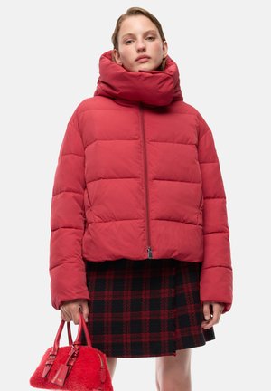 Donna che indossa una giacca a vento rossa e una gonna a quadri nera e rossa, che tiene in mano una borsa rossa su sfondo bianco.