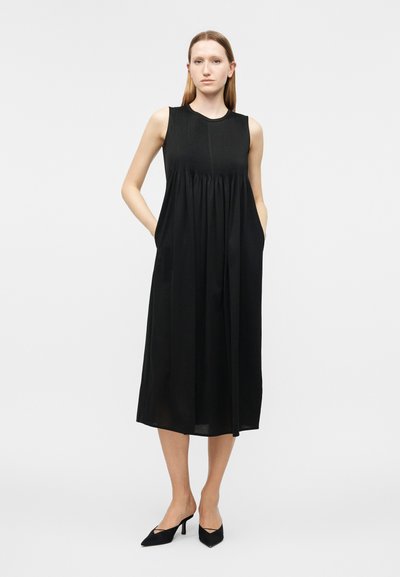 Γυναίκα που στέκεται, φοράει μαύρο μίντι φόρεμα χωρίς μανίκια με πλισέ μπούστο και τσέπες, συνδυασμένο με μαύρες μυτερές γόβες, σε απλό φόντο.