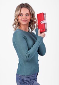 Red wallet with a polka dot accent, smooth texture, and a snap closure, held against a white background by a person in a teal long-sleeve shirt.