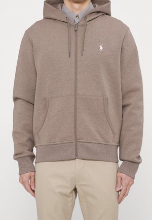 Man draagt een beige zip-up hoodie met trekkoorden en een kangoeroezak, gecombineerd met lichte kaki broek en een grijs overhemd met kraag eronder.