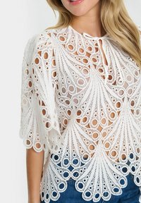 Blusa bianca in pizzo con intricati design a intaglio, scollatura rotonda con legatura e maniche a sbuffo, abbinata a jeans blu.