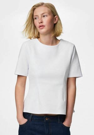 Blond kvinne iført en enkel hvit kortermet topp og mørkeblå jeans, stående med hendene i lommene, ser mot siden.