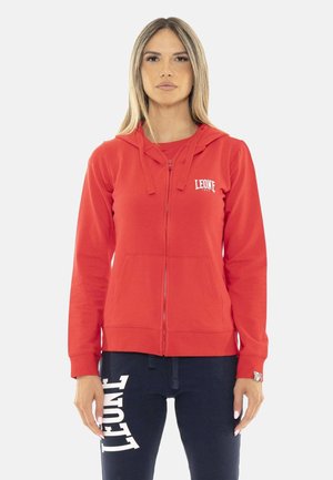 Vrouw draagt een rode hoodie met ritssluiting met "LEONE"-logo en donkerblauwe broek met grote "LEONE"-tekst op de linker dij, staand tegen een witte achtergrond.