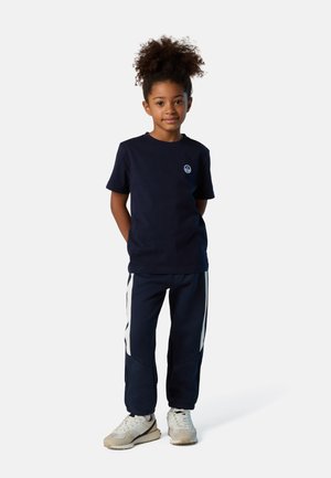 Camiseta navy con un pequeño logo en el pecho, combinada con pantalones de chándal navy que tienen franjas laterales blancas. El modelo lleva zapatillas ligeras.