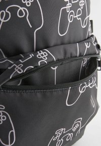 Schwarzer Stoff-Rucksack mit einem wiederholenden weißen Muster von Gaming-Controllern. Enthält eine vordere Reißverschlusstasche und strapazierfähige Nähte.