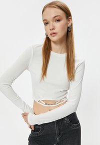 Top corto blanco acanalado de mangas largas, con un corte en la cintura adornado con una cadena plateada; combinado con unos jeans negros de cintura alta.