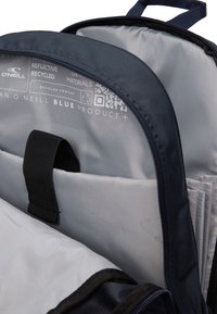 Granatowy plecak z szarym wnętrzem, posiadający wiele przegród, regulowane paski oraz etykiety recyklingowe. Widoczny zamek błyskawiczny.