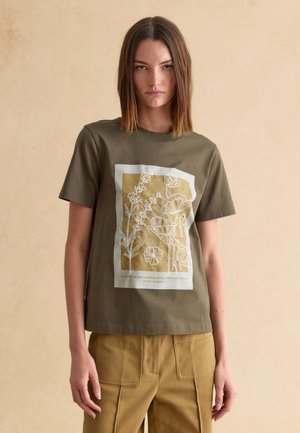 Jonge vrouw met een olijfgroen T-shirt met wit en mosterdkleurig bloemmotief, gecombineerd met mosterdkleurige broek, staande tegen een beige achtergrond.