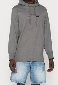Sweat à capuche gris avec cordons, présentant un petit motif de texte imprimé. Associé à un short en denim bleu délavé avec un patch.