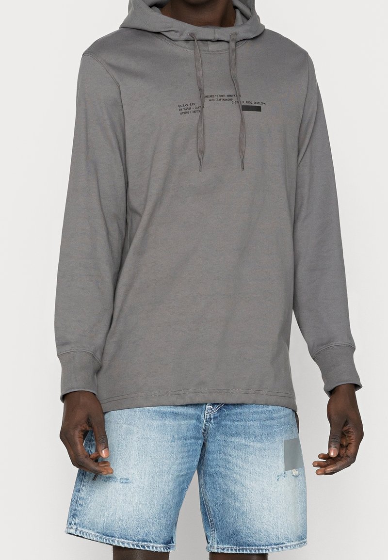 Sweat à capuche gris avec cordons, présentant un petit motif de texte imprimé. Associé à un short en denim bleu délavé avec un patch.