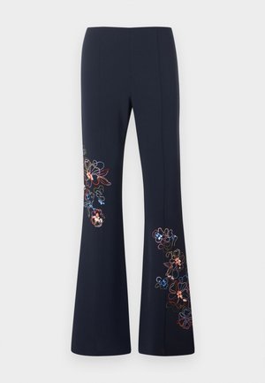 Kobieta stoi ze skrzyżowanymi ramionami, ubrana w jasnoniebieski top bez rękawów i czarne spodnie o szerokich nogawkach z haftowanymi kwiatowymi wzorami.