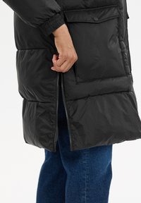 Svart puffjacka med dragkedja framtill, elastiska ärmslut och tryckknappar på fickorna. Tyget ser blanka ut med en slät yta.