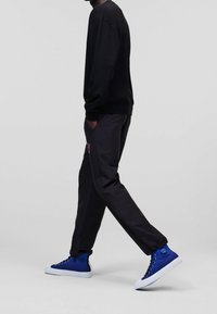 Felpa nera con polsini a costine, abbinata a pantaloni scuri con un dettaglio ricamato rosa, e sneakers alte blu con suole bianche.