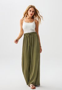 Pantaloni a gamba larga color oliva con tessuto strutturato, vita con coulisse, abbinati a un top bianco aderente e sandali marroni.