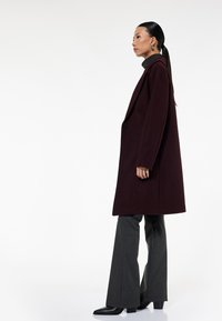 Bordeaux uldfrakke med reverskrave, enkelt knaplukning og to sidelommer, parret med grå flagrende bukser og sorte sko.