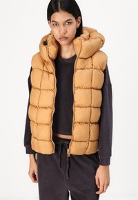Gilet imbottito di colore marrone chiaro, con sezioni trapuntate, colletto alto e zip a tutta lunghezza. Indossato sopra un maglione scuro a coste.