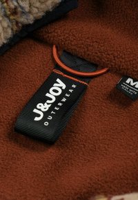 Bruin fleece stof met een gestructureerd oppervlak, voorzien van een opvallend zwart label met de tekst "J&JOY OUTERWEAR" en een oranje accentlus.