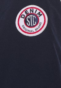 Marinblått tyg med en texturerad yta, med en cirkulär röd och vit broderad lapp som säger "DENIM" och "ORIGINAL BRAND."