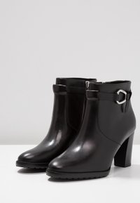 Botas de tobillo de cuero negro con un tacón alto, cierre de cremallera y una correa decorativa con un hebilla de plata. Textura lisa y punta redondeada.
