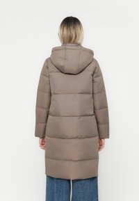 Manteau matelassé marron avec capuche, présentant des sections matelassées, des manches longues et une coupe droite, porté sur un jean en denim à jambe large.