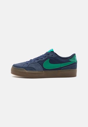 Baskets basses bleu marine avec un swoosh Nike vert, languette de talon bleu sarcelle et semelle en caoutchouc gomme, montrées de l’intérieur sur fond blanc.