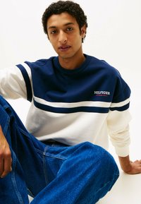 Felpa navy e bianca con strisce orizzontali, presenta il logo "HILFIGER". Indossata con jeans in denim blu, tessuto testurizzato, design a girocollo.