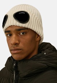 Beanie bianco lavorato a maglia con occhiali neri distintivi sopra, texture a costine e un accento con logo. Il modello indossa una giacca nera con zip.