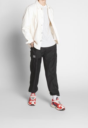 Chaqueta de denim blanca, camiseta blanca a rayas, pantalones cargo negros y zapatillas rojas/blancas con detalles azules. Atuendo casual con texturas variadas.