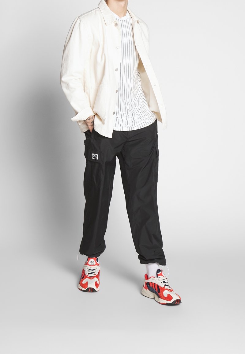 Valkoinen farkkutakki, raidallinen valkoinen t-paita, mustat cargo-housut ja punaiset/valkoiset lenkkarit sinisillä yksityiskohdilla. Rentoa asua vaihtelevilla tekstuureilla.