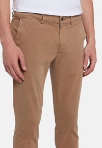 Pantaloni in cotone marrone con una texture liscia, design slim fit, tasche laterali e chiusura con zip e bottone in metallo.