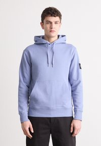Hanorac bleu deschis, fabricat dintr-un material moale, având o buzunare frontală, șnururi, manșete ribbate și un detaliu cu patch pe lateral.