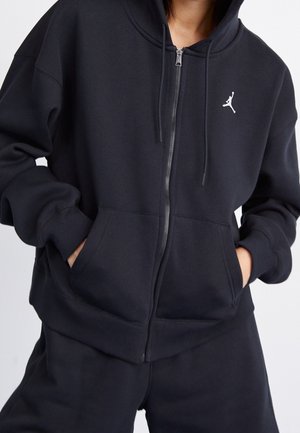 Person trägt einen schwarzen Kapuzenpullover mit Reißverschluss und weißem Jumpman-Logo, die Hände in den Fronttaschen, vor einem einfarbigen Hintergrund.