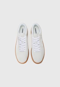 White suede sneakers with a gum rubber sole, featuring white laces and branding on the tongue. Smooth texture and rounded toe design.