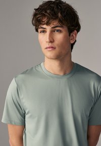Camiseta atlética verde claro con un patrón texturizado, cuello redondo, mangas cortas y tela suave. Acercamiento de la parte superior del cuerpo.