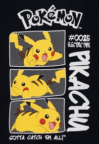 Melnas krekls ar trim Pikachu ilustrācijām dzeltenā un melnā krāsu shēmā, ar punktiņu rakstiem un tekstu "Pokémon" un "Pikachu."