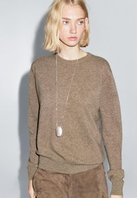 Maglione di lana marrone con scollatura a girocollo e polsini a coste, abbinato a pantaloni in camoscio marrone e un pendente argentato su una lunga catenina.