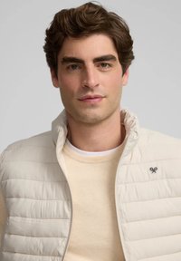 Chaleco de plumas ligero con costuras horizontales, cuello alto y pequeño logo, superpuesto a un suave suéter beige de cuello redondo.