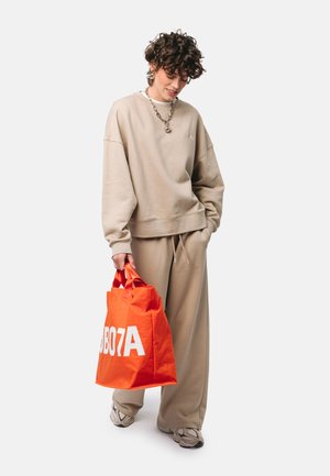 Osoba w beżowej, oversize bluzie i spodniach trzyma jasnopomarańczową torbę na ramię z białym napisem, patrzy w dół i się uśmiecha, stoi na białym tle.