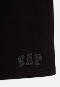 Melna kokvilnas t-kreklu ar teksturētu adījumu, ar pelēku izšūtu "GAP" logo kreisajā apakšējā stūrī un šūtām apmalēm.