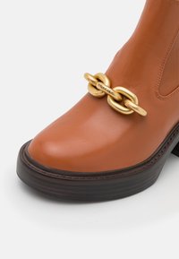 Botte en cuir couleur fauve avec semelle marron foncé, présentant un grand détail de chaîne en or gravé "Coach" à l'avant.