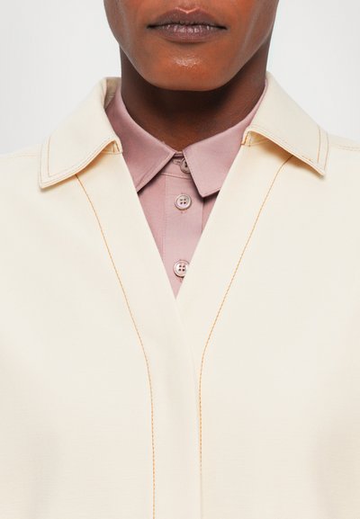 Κρεμ σακάκι με φαρδύ γιακά, που διαθέτει αντίθεση πορτοκαλί ραφές; συνδυασμένο με ένα ανοιχτό ροζ πουκάμισο από κάτω.