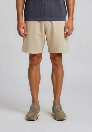 Hombre con pantalones cortos beige con logo blanco en la pierna izquierda, camiseta gris acanalada y zapatillas color topo, de pie contra un fondo liso.