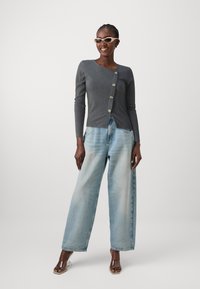 Haut gris à manches longues en côte avec détail de boutons, associé à un jean large bleu clair. Des sandales à talons transparents complètent la tenue.
